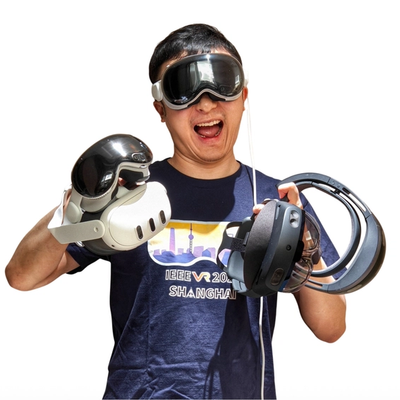 Man wearing a VR headset and holding four more VR headsets in his hands. Has excited expression, smiling with an open mouth. Short dark hair. 